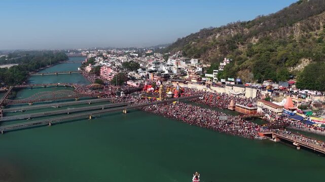 Push in drone view of Har Ki Pauri in Haridwar showing crowded Ganga ghat, bridges, and temples. Devotees gather along riverbanks performing rituals in a vibrant spiritual atmosphere.