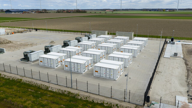 Dronten, Netherlands - 04 April 2026: Aerial view of the Olsterdwarspad battery energy storage system facility with rows of white containers and wind turbines under a cloudy sky.