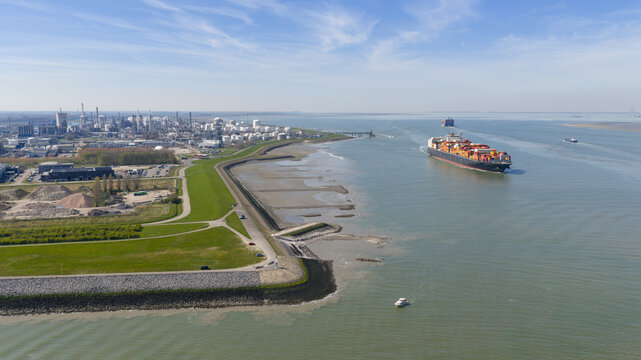 Terneuzen, Netherlands - 08 April 2026: Aerial view of a large container ship navigating the Western Scheldt river past the industrial port area and green dikes under a clear blue sky.