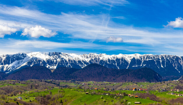 A gorgeous landscape with the snow-covered Piatra Craiului mountains in the background and the houses of the village of Pestera with green fields in the foreground in spring