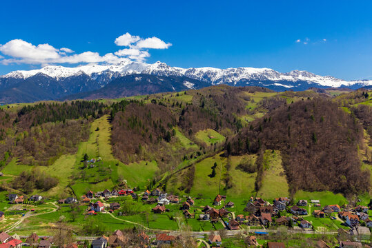 Picturesque landscape of the Bucegi Mountains with snow-capped peaks in the background and the village of Moieciu between green hills in the foreground
