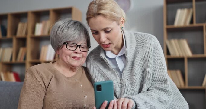 Senior woman and daughter video chat by phone. On a sofa the younger guides her mother through apps, makes calls, and practices safe online use. Concept intergenerational digital care at home.