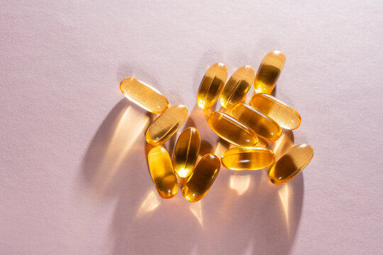 Golden liquid gel capsules on purple background. Translucent amber fish oil or vitamin softgels scattered on matte lilac surface with dramatic shadows. Minimalist healthy lifestyle concept