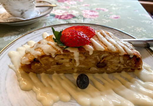 A delicious slice of fresh apple strudel served on a white plate with creamy vanilla sauce, topped with sliced strawberries and a blueberry