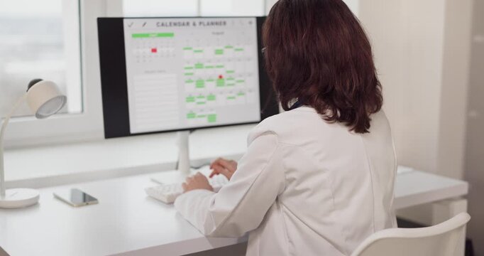 Doctor woman working at computer in office. In a clean clinic she reviews a calendar on the monitor, managing scheduling and records at a tidy desk. Healthcare workflow and productivity concept.