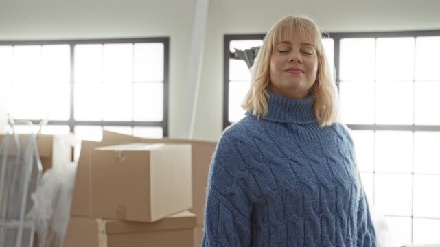 Woman standing among cardboard moving boxes, wearing blue turtleneck sweater with eyes closed and slight smile in studio; new beginning contentment.