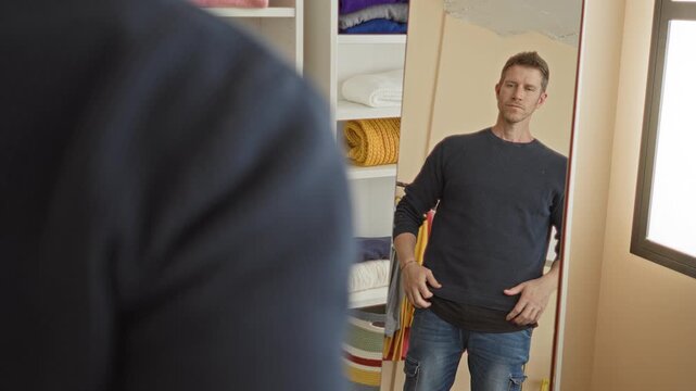 Man adjusts navy sweater hem, rolls sleeve and checks fit in full length mirror beside shelves of folded blankets in building; introspection.