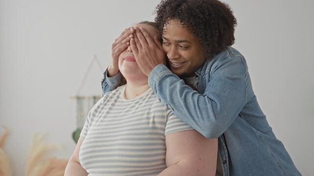 Woman smiles as man covers her eyes with his hands and hugs her in a studio; intimacy warmth joy playful.
