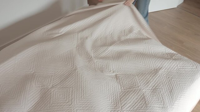 Woman spreads light coverlet over bed in bedroom interior. Bed making, home routine and domestic lifestyle concepts