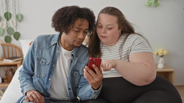Man and plus size woman sit close on bed and point finger to red smartphone, showing screen beside bedside table and vase in bedroom; casual affection.