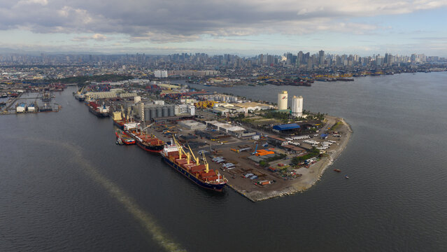Manila, Philippines - 01 March 2026: Aerial view of Manila Multipurpose Terminal with cargo ships docked at the pier and the city skyline under a cloudy sky.