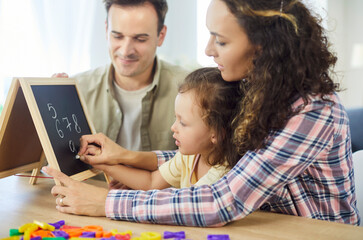 Family parents guide daughter writing numbers and alphabet. At home, child girl learning on a chalkboard as parents guide, making education and homework easier. Supportive education and development. © Studio Romantic