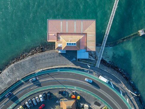 High-angle view of a coastal road curving around a harbor building. Cars and parked vehicles are visible. A pedestrian bridge extends into the water. TAMAKI DRIVE, OKAHU BAY, AUCKLAND, NEW ZEALAND