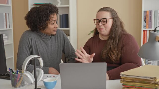 Man and woman at laptop, man points finger to laptop screen in building office, woman shows open hand gesture; collaboration concentration.