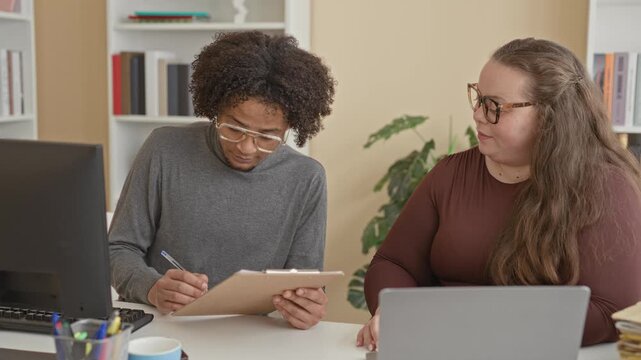 Man and woman at desk exchange a clipboard and shake hands in an office meeting with laptop and folders on table; teamwork trust.