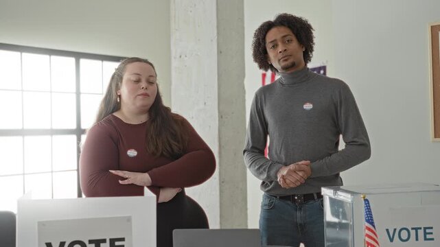 Man and woman stand by a transparent ballot box, man with hands clasped and both wearing i voted stickers inside a polling place building; civic duty pride.