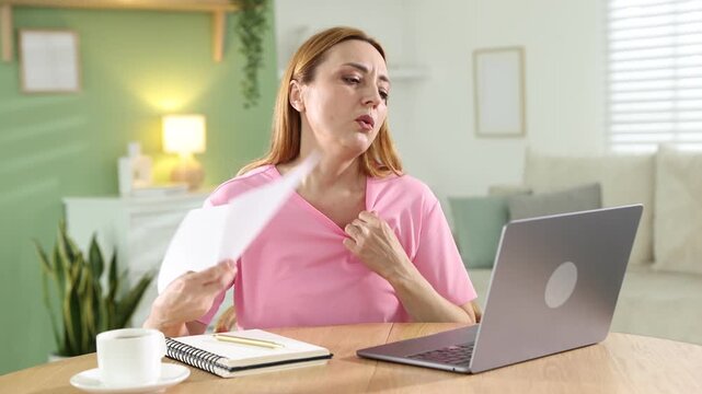 Menopause. Woman waving paper sheet to cool herself while working on laptop at table indoors