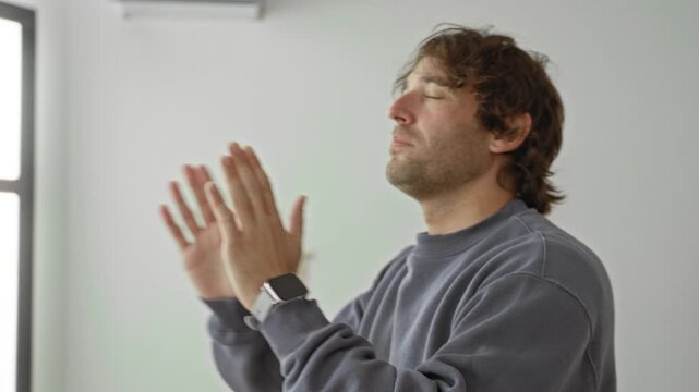 Man with hands pressed together in prayer pose, eyes closed and smartwatch visible, standing against macrame wall in studio; calm meditation.