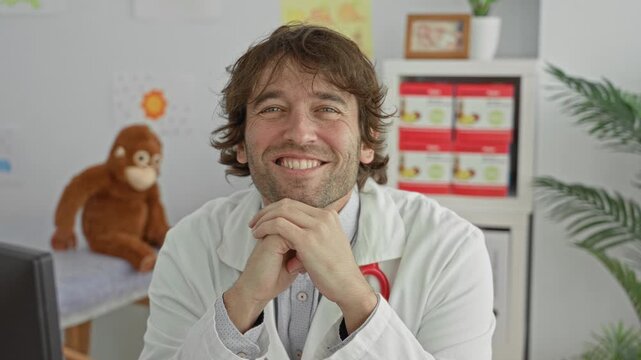 Man doctor with stethoscope, hands clasped and smiling in pediatric clinic with stuffed monkey and exam table; comfort trust.