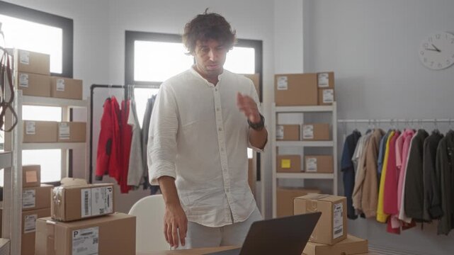 Man holding forehead beside laptop and stacked shipping boxes and clothing rack in studio while checking orders; stress ecommerce operations.