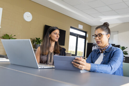 Diverse female coworkers discussing project at office as one holding tablet case other using laptop