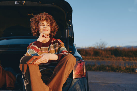Woman lifestyle portrait in a parked car at sunset, smile on her face, curly hair, outdoors countryside road view, chin on hand inside the car, warm light, relax close view
