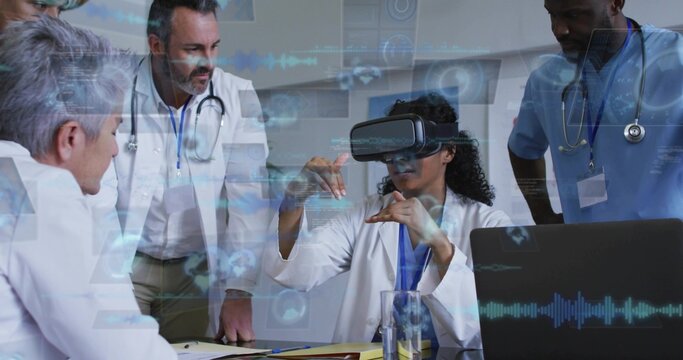 Wearing VR headset clinician in lab coat gesturing at medical table, team watching laptop holograms