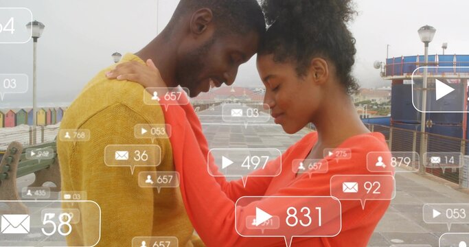 Pressing foreheads, couple wearing mustard and coral sweaters on seafront with notification badges