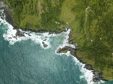 Aerial view of Banyutibo Water Fall cascading into the turquoise ocean with white waves crashing against rocky cliffs and lush green hills in Girisubo, Special Region of Yogyakarta, Indonesia.