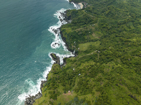 Aerial view of Banyu Tibo Waterfall cascading from lush green cliffs into the turquoise ocean waves along the rugged coastline of Girisubo, Special Region of Yogyakarta, Indonesia.