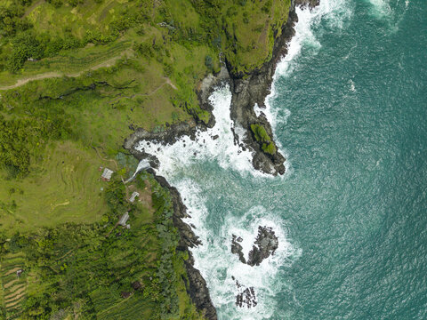 Aerial view of Banyutibo Water Fall cascading into the turquoise ocean with waves crashing against dark rocks and lush green cliffs in Girisubo, Special Region of Yogyakarta, Indonesia.