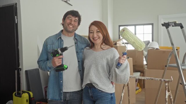 Man holds drill and woman holds paint roller smiling amid packed moving boxes, ladder and hard hat while unpacking in building; home renovation joy.