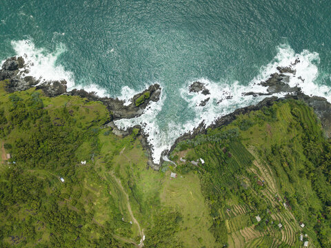 Aerial view of Banyutibo Water Fall cascading over dark cliffs into the turquoise ocean waves, surrounded by lush green terraced fields Girisubo, Special Region of Yogyakarta, Indonesia.
