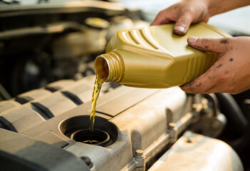 Obraz premium A person with dirty hands pours fresh golden oil from a yellow bottle into a car engine, performing vehicle maintenance.