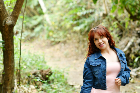 Mujer latina de pie sobre un camino de tierra en medio de un ambiente natural, usando un vestido rosado y chaqueta azul. Toma horizontal, espacio para texto al lado izquierdo.