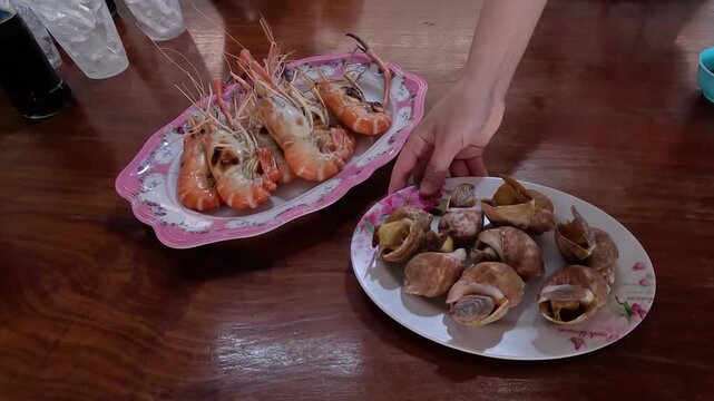 A close up cinematic motion of someone setting down plates of freshly cooked grilled prawns, boiled spotted babylon snails, and various dipping sauces on a wooden table at a seafood restaurant.