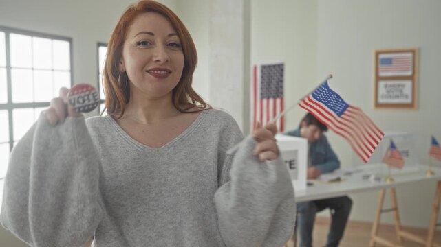 Woman holds vote button and waves american flag inside a building voting area beside a ballot box and voting booth; civic pride.