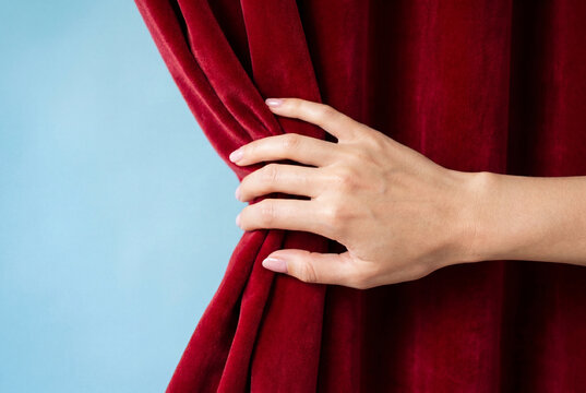 Elegant hand gently pulling back a heavy red velvet curtain to reveal a light blue open space