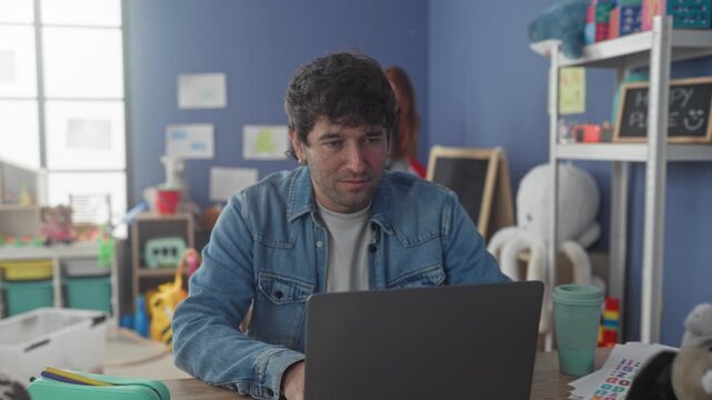 Man smiling while typing on laptop in preschool classroom with woman teacher tidying toys in background; family balance joy.