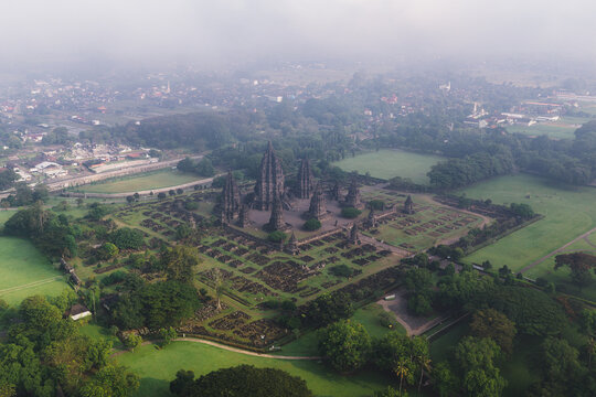 Aerial view of Prambanan Temple, the ancient Hindu temple complex with towering stone spires surrounded by green lawns and misty haze in Kecamatan Prambanan, Daerah Istimewa Yogyakarta, Indonesia.