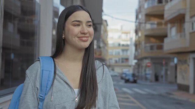 Young hispanic woman wearing a blue backpack smiles while looking to the right on a city street with apartment buildings; optimism.