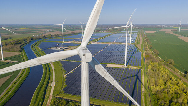 Aerial view of Zonnepark Eekerpolder featuring wind turbines and vast solar panel arrays next to a canal in Scheemda, Groningen, Netherlands.