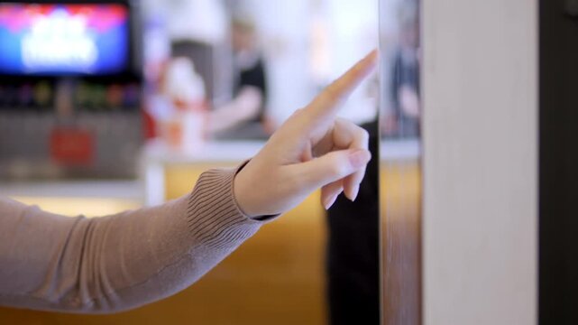 Hand points finger at touch screen. Background shows blurred cafe interior. Action shows interaction with digital interface. Person wears sweater, hand gestures precisely