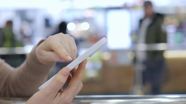 Hands hold white smartphone, thumbs scroll screen. Background blurred, people move behind glass. Indoor setting with warm lighting. Focus on device interaction, natural movement