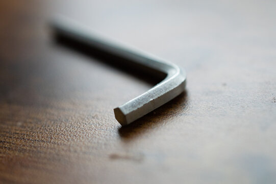 Close-up macro shot of a metallic silver L-shaped hex key Allen wrench lying on a textured wooden surface, highlighting the essential tool for assembly and repair tasks with shallow depth of field.