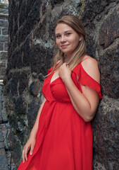 Fototapeta premium Smiling young woman in red dress poses against rugged stone wall on a cobblestone street