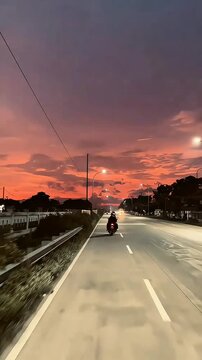 A lone motorcyclist rides down a long road as the sky explodes in a beautiful sunset of pinks, purples, and oranges, creating a serene atmosphere.