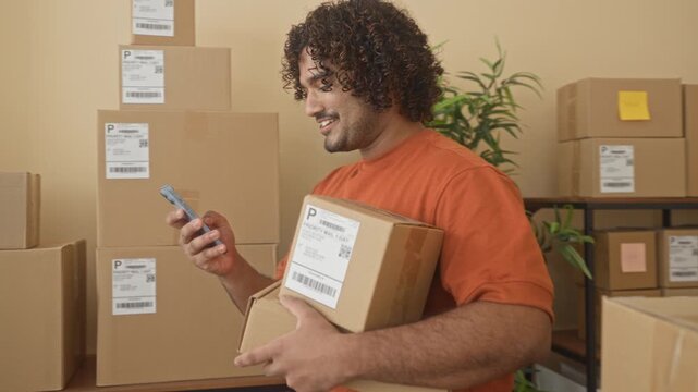 Man holding cardboard box with smartphone in hand, smiling amid stacked parcels, shipping labels and a potted plant on shelves inside a building depot; small business optimism.