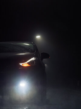 A dark car sits parked on a wet road at night. Its bright headlights, daytime running lights, and fog lights cut through the heavy mist.