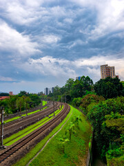 Obraz premium Curving railway tracks pass through a vibrant green embankment beside dense city vegetation in Medellin, Antioquia, with tall buildings in the distance under a dramatic cloudy sky.
